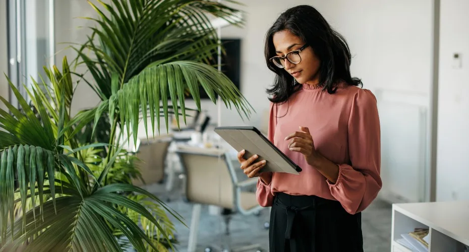 A woman wearing glasses is focused on using a tablet while seated at her office desk.