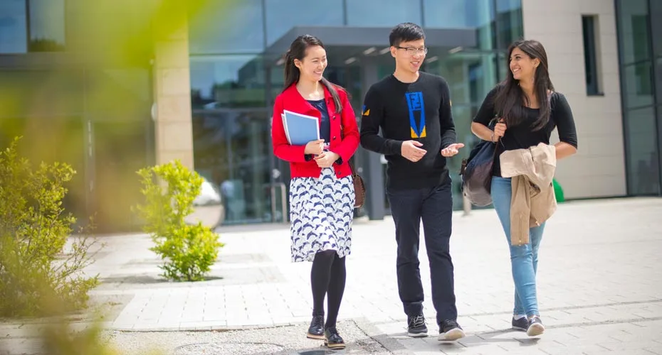 Three students walking on a sidewalk outside building one at the university of exeter.